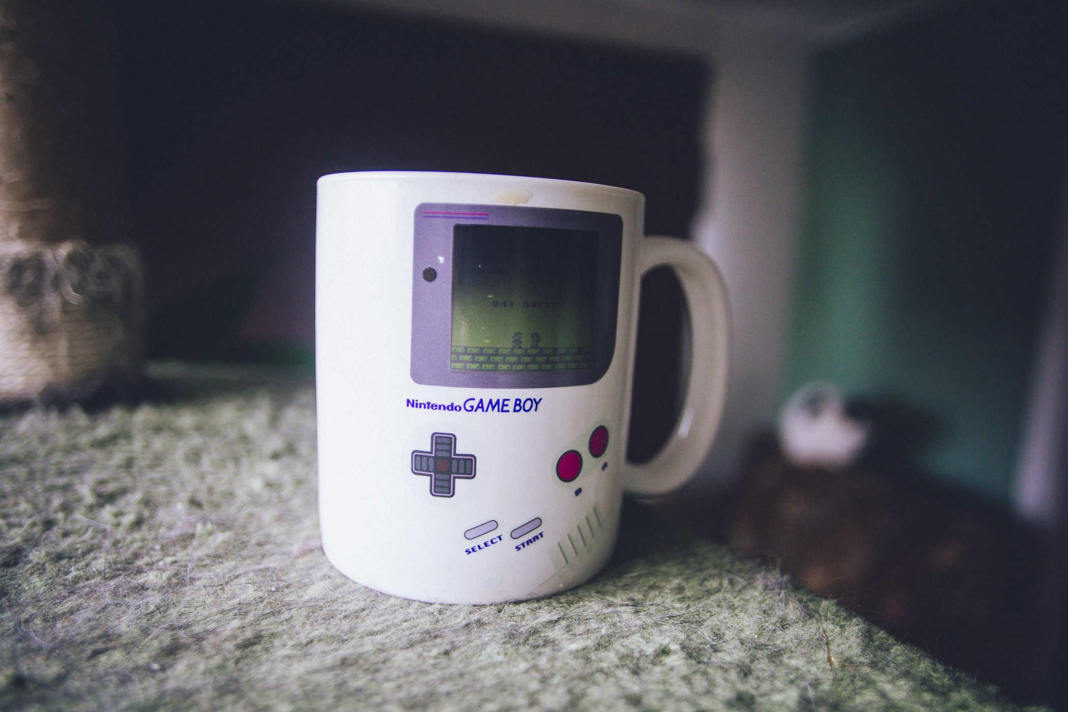 A white mug with the face of a Nintendo Game Boy on it that is heat sensitive and shows a game of Super Mario land on it when heated; a white cat rests in the background