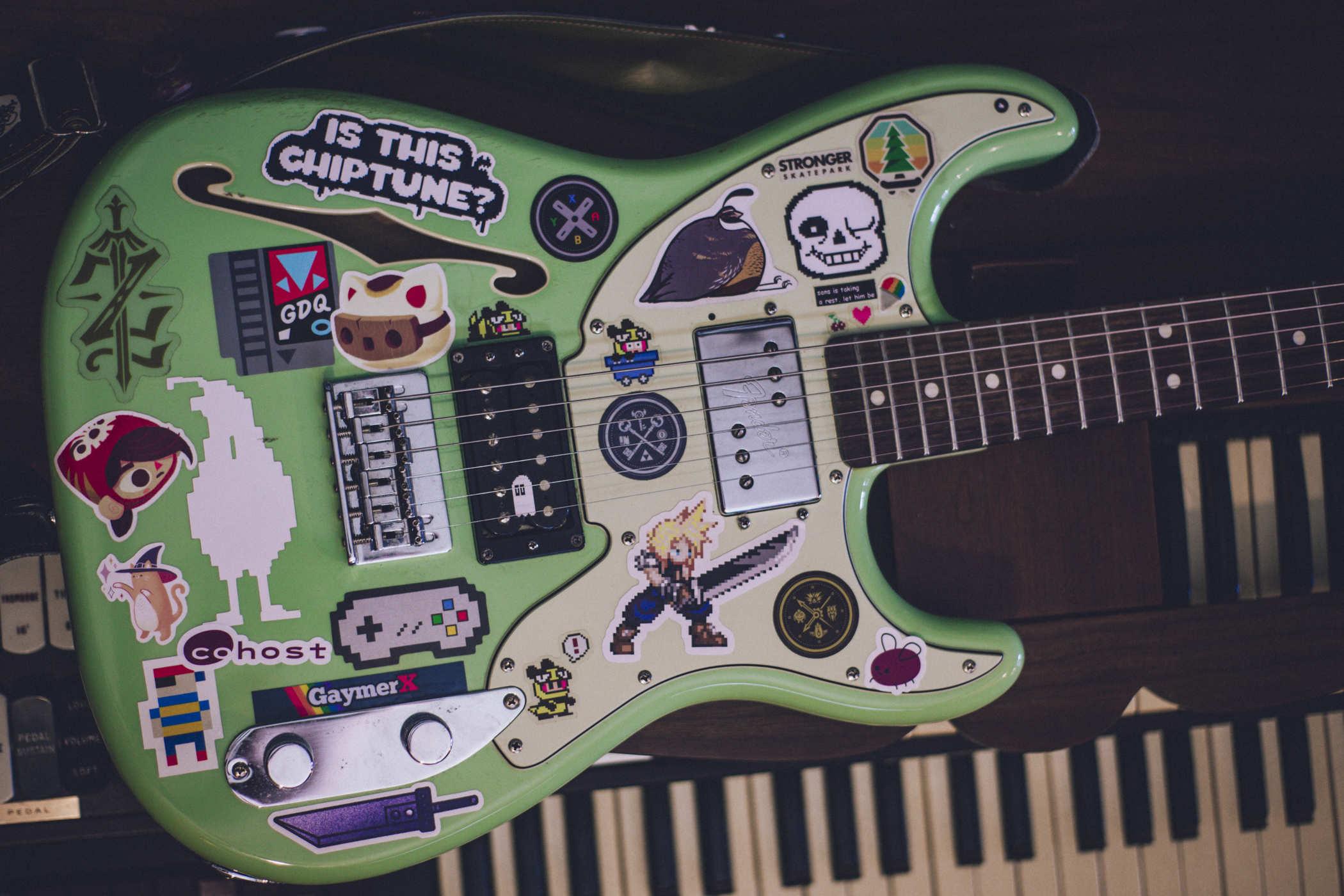 Photo of a seafoam green guitar covered in stickers ranging from 'IS THIS CHIPTUNE?' to video game contollers, but prominent is the character Cloud Strife, a spikey-haired blonde guy with a massive sword