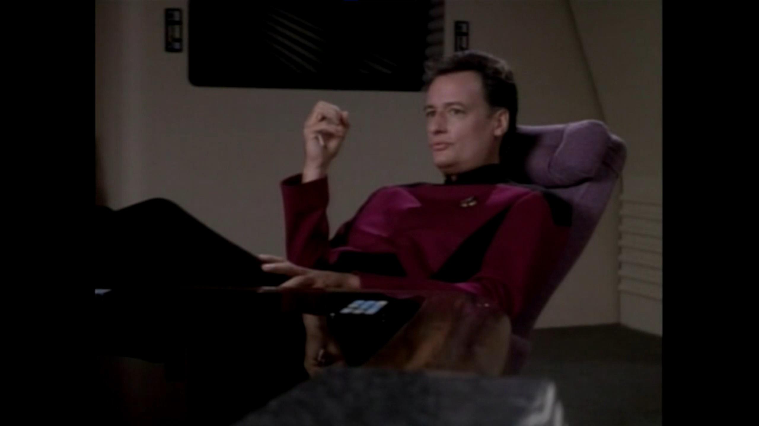 Screenshot of a Star Trek TNG episode where John de Lancie as 'Q' sits in a chair in a red command starfeleet uniform and appears to be holding up an invisible ball