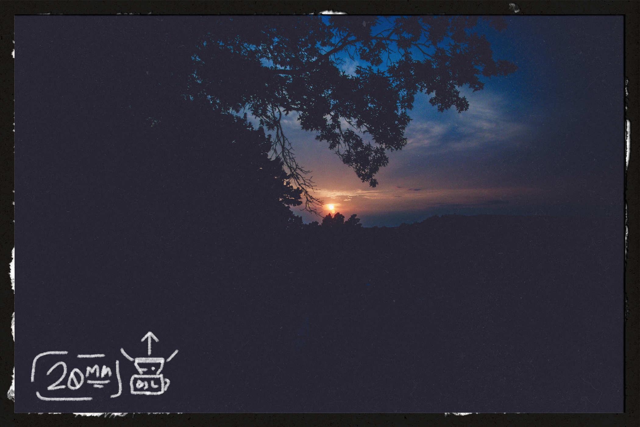 The sun is a tiny dot in the distance and a tree in silhouette takes up most of the frame; handwritten in the lower left is '20mm' and a drawing of a camera
