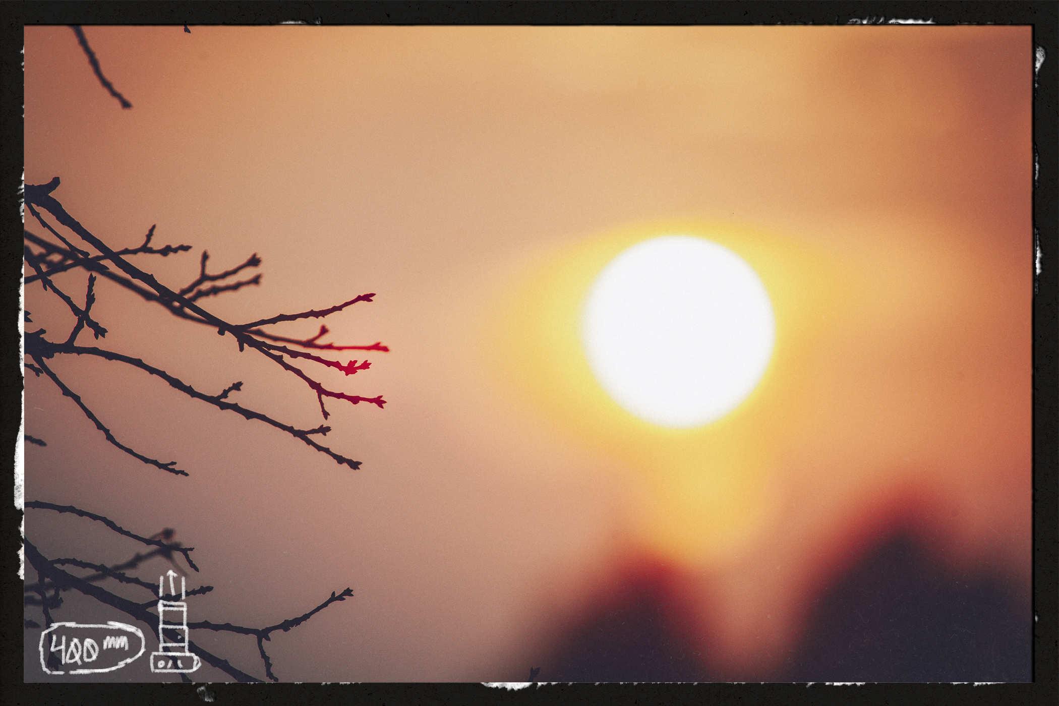Photo of the sun with some tree branch twigs in silhouette in the foreground; handwritten in the lower left is '400mm' and a drawing of a camer 