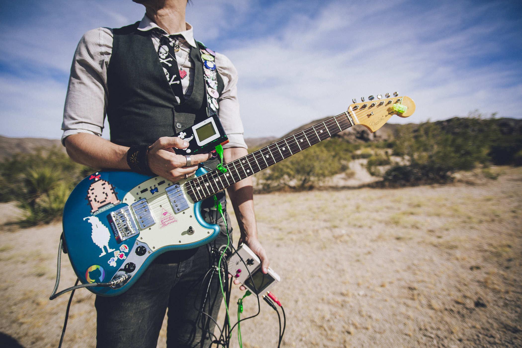 Photo of a person standing in the middle of Joshua Tree National Park wearing a guitar and holding up multiple Nintendo Gameboys