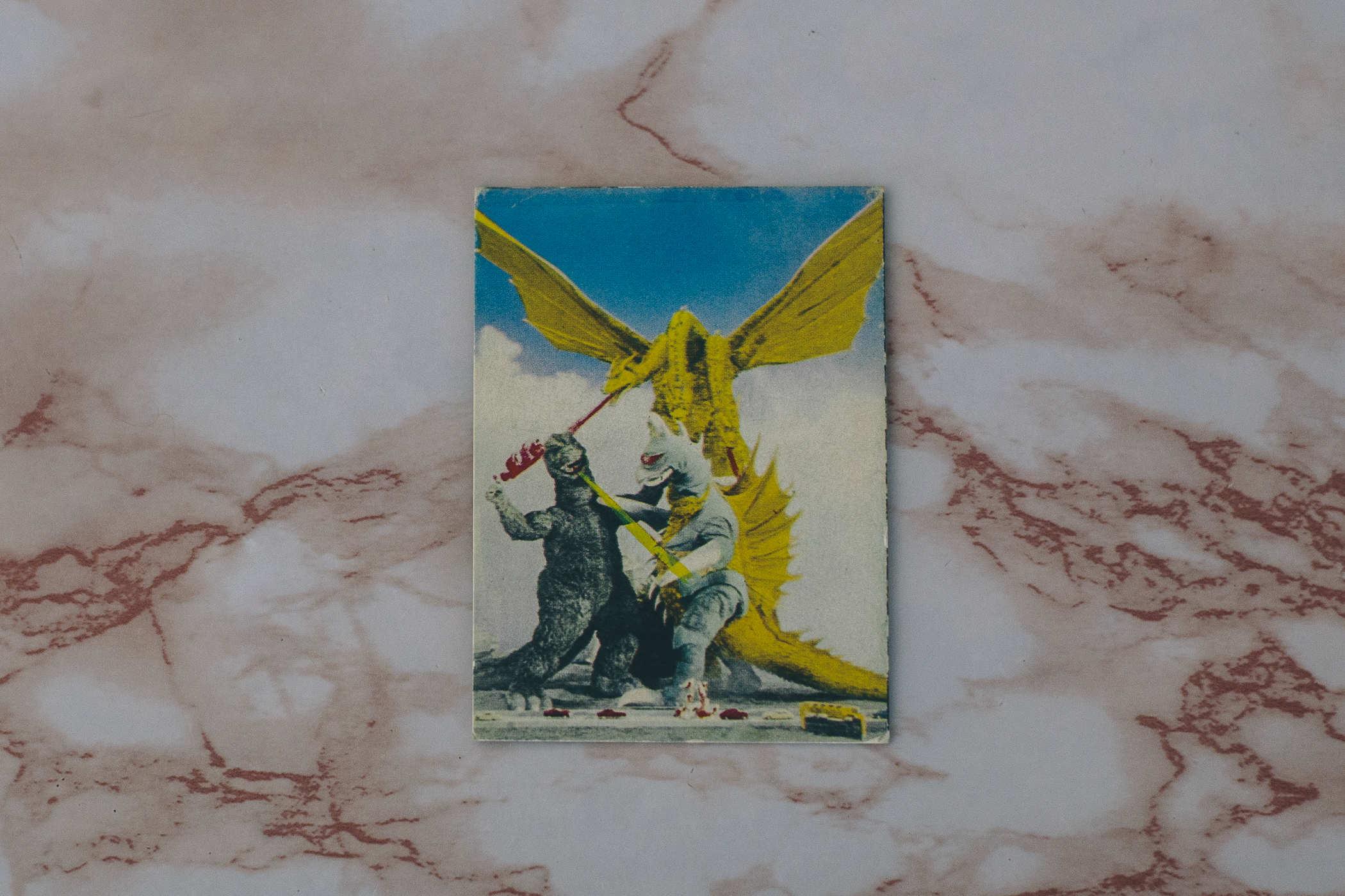 Photo of a small card on a pink countertop; the card has an image of Godzilla fighting a bird-beaked biped monster (Gigan) and a large golden three-headed dragon (Ghidorah) 
