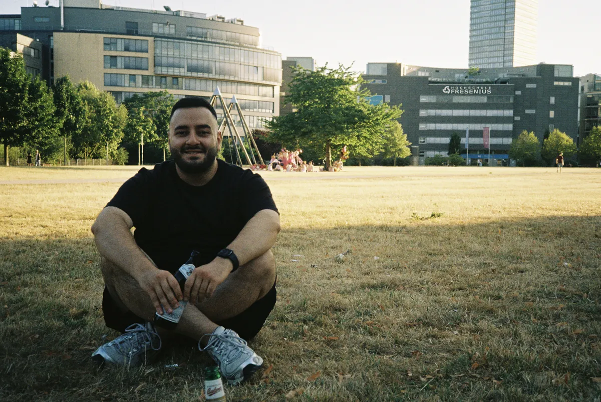 An adult man with a dark beard and short hair, named Dastan, sits cross-legged on a dry grassy field in a park during a sunny afternoon. He is wearing a black t-shirt, dark shorts, grey sneakers, and a watch on his left wrist. He is smiling warmly at the camera while holding a beverage bottle in his hand, with another green bottle resting on the grass in the foreground.
In the background, there is a sprawling green field with trees, some people gathered in the distance, and prominent city buildings, including one with 