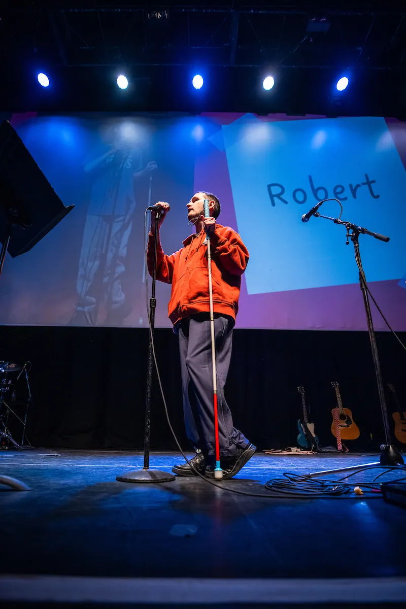 Robert Kingett speaking on stage with one hand on the microphone and looking to the side, as seen from the front row, with his name projected on the wall behind him