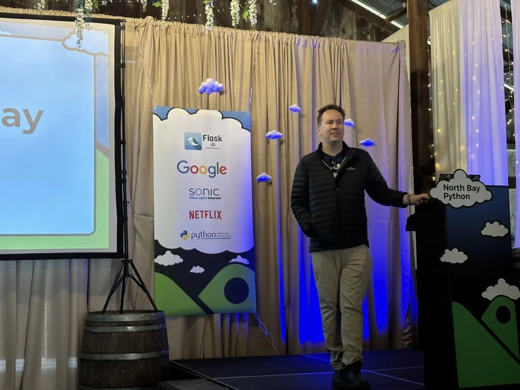 Christopher standing on stage behind the NBPy logo, introducing the conference, with a sponsor banner with the Flask logo at the top