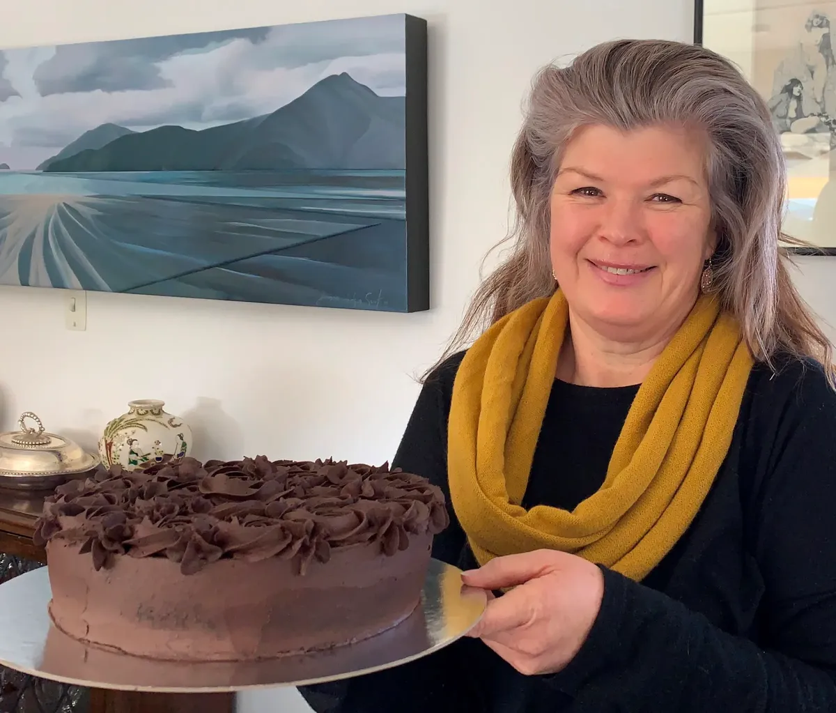 Jude, wearing a mustard yellow scarf and indigo top, smiling as she holds up her Marlborough Mud Cake.