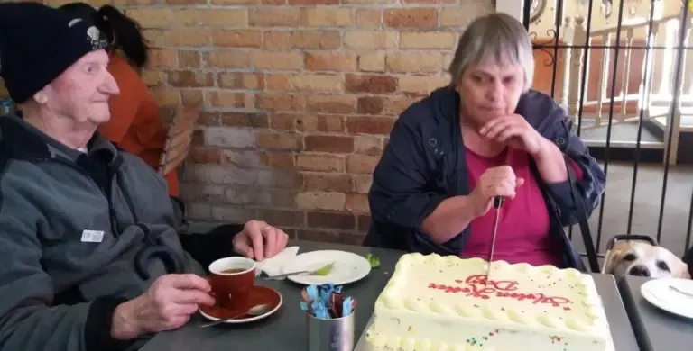 Joan, a Waikato deafblind network member, cutting the cake, with another network member and the top of a hopeful-looking guide dog's head sharing the table.