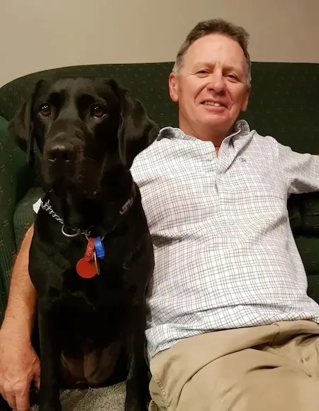 David Wilson sits on a couch with his guide dog, a black Labrador named Cayla.