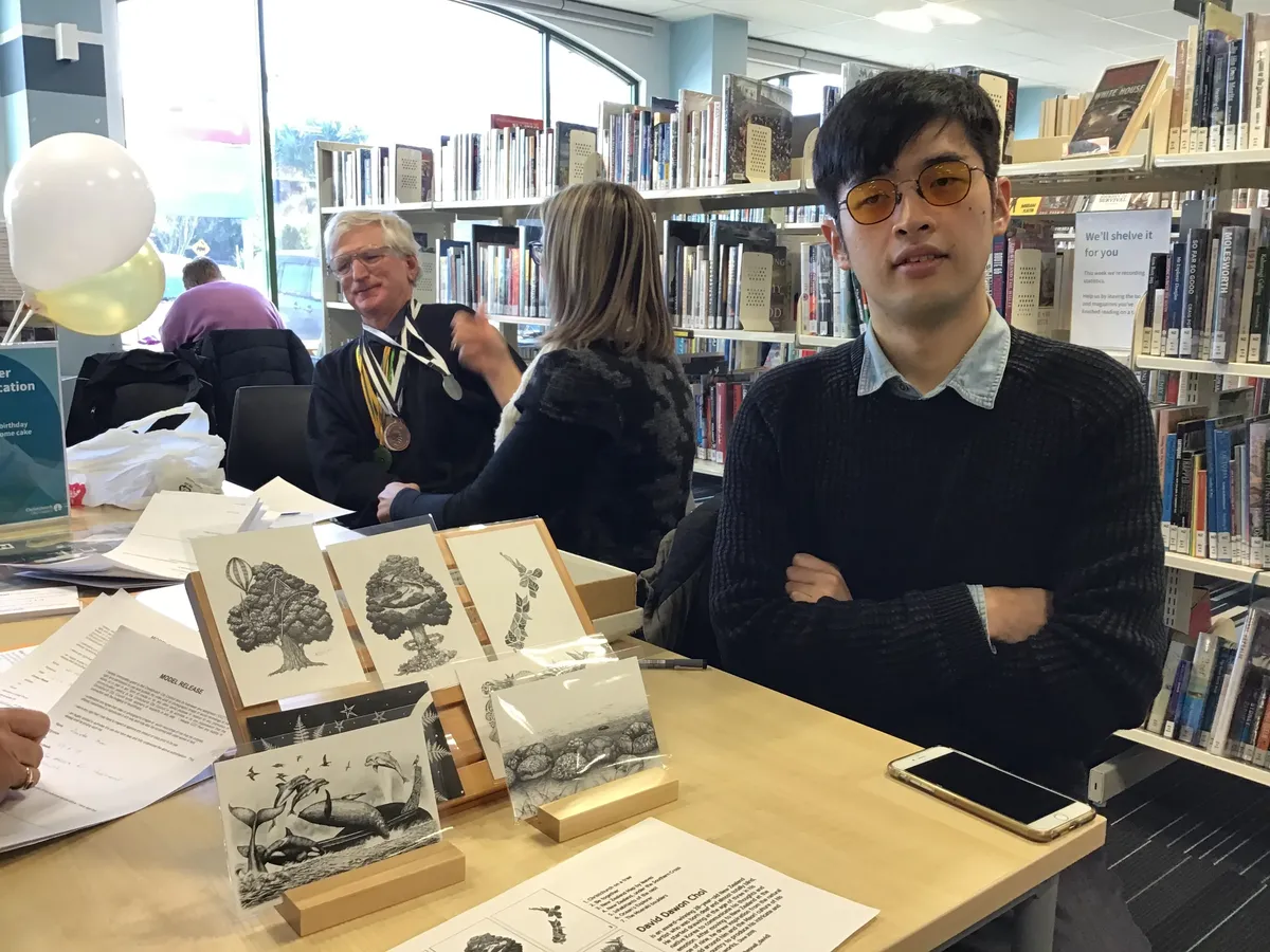 A deafblind artist looks at the camera, arms crossed, next to examples of his sketches.
