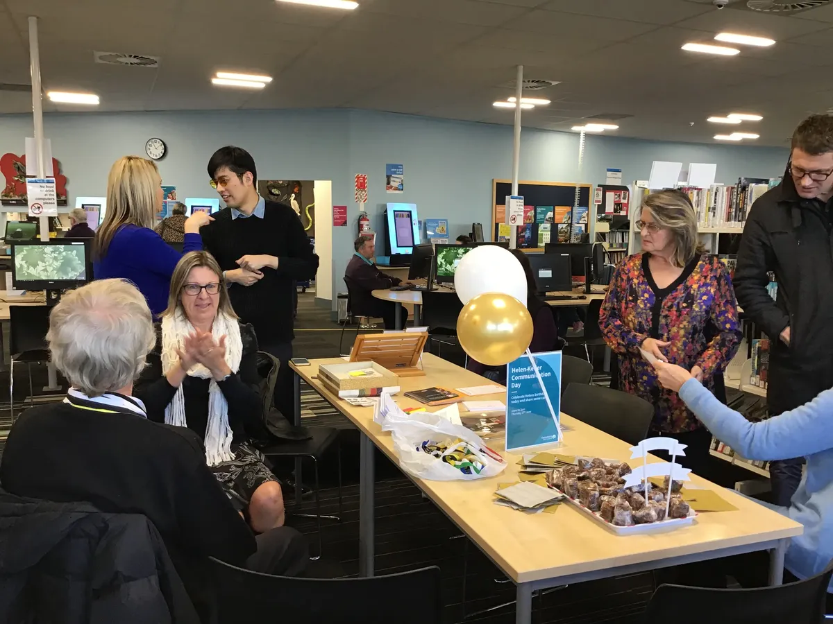 Bowler and artist around table of snacks and balloons. They're talking with iSign interpreters, with Kaye Ellison looking on