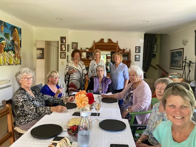 9 women are gathered at a dining table formally set for 10 people. The plates are charcoal on white table cloth, and dahlias in fire colours are in a crystal vase on the table. The women are smiling and holding flutes of bubbles. There is a kauri dresser in the background and photographs on the walls.