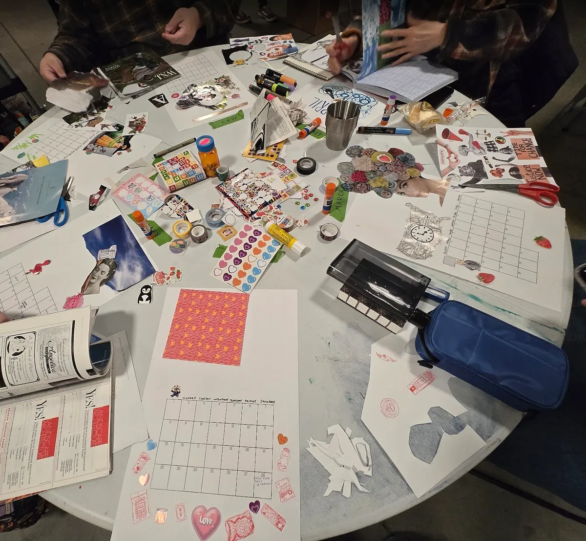 Photo of people collaging on the calendar pages, the table is messy with cut up magazines