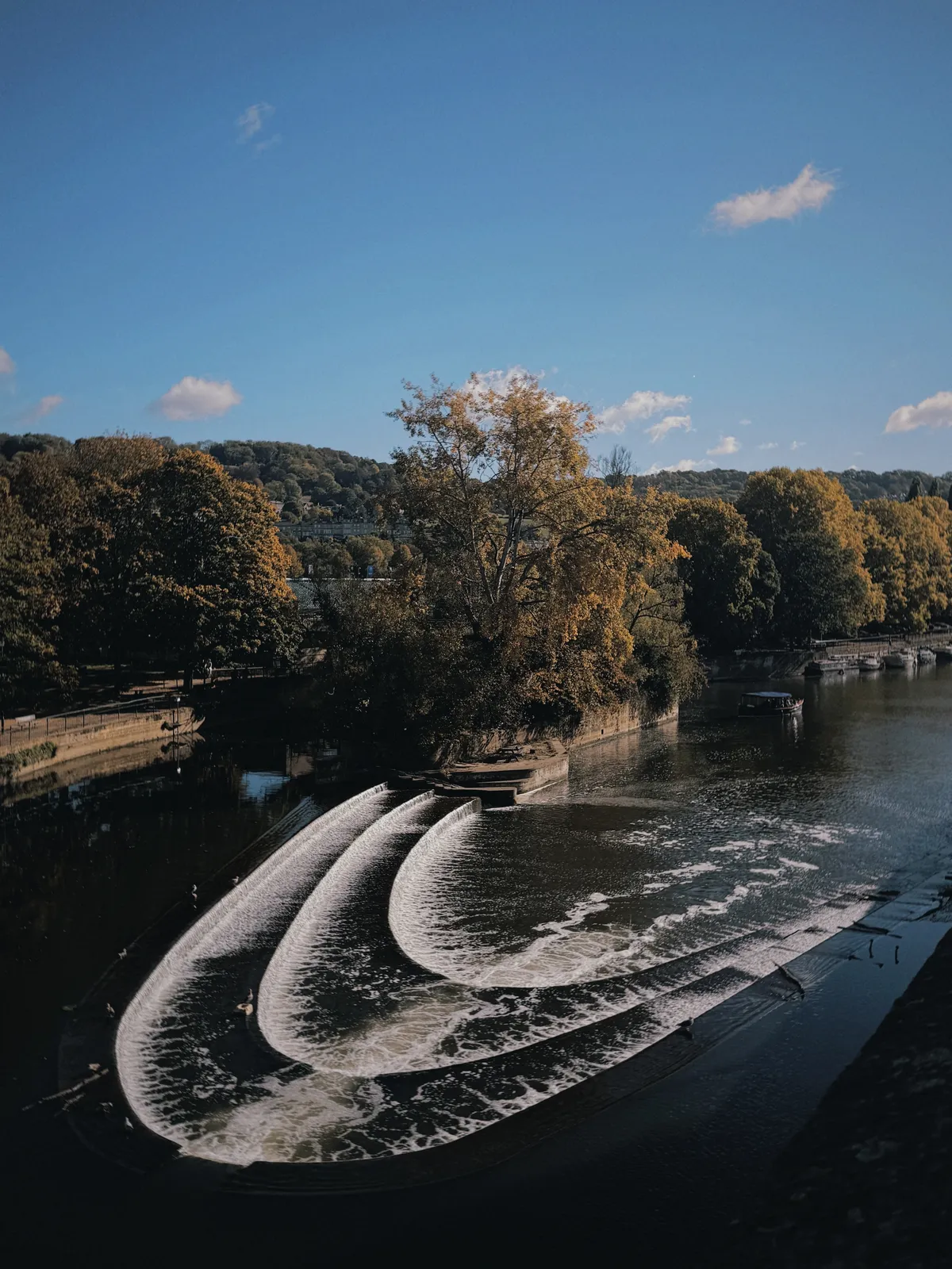 Pulteney Weir in Bath on a sunny autumn day, water spilling over its curved, stepped tiers into the River Avon