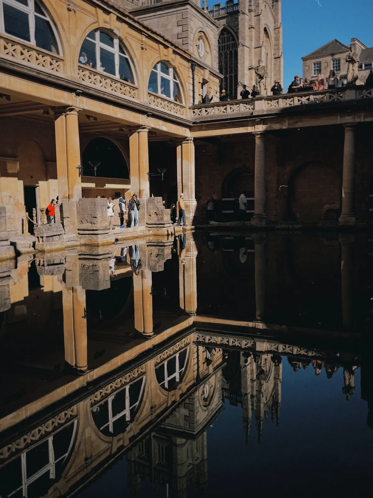 The Great Bath at the Roman Baths, arched windows and the tower of Bath Abbey mirrored in the still, dark green water, with visitors standing along the terrace above