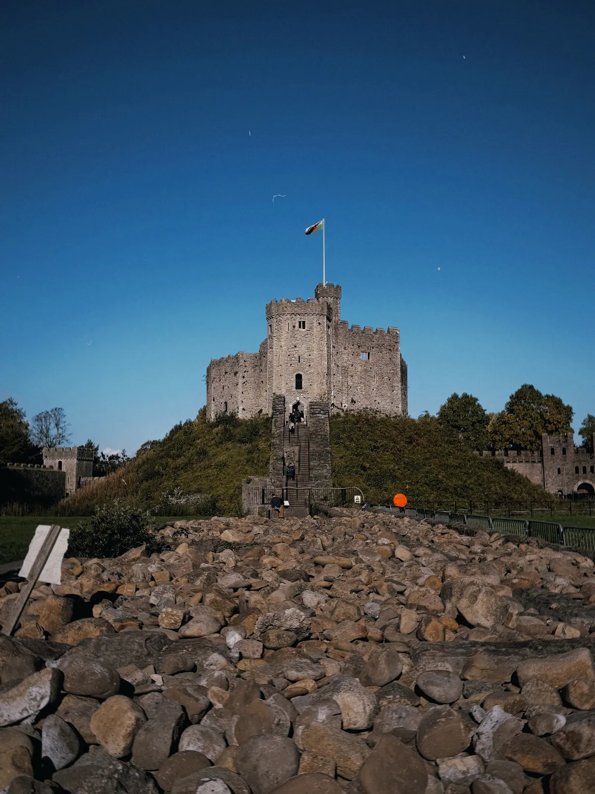 The Norman keep of Cardiff Castle on its grassy motte under a deep blue sky, a Welsh flag flying from the top and a stone stairway leading up, with a bed of large pebbles in the foreground