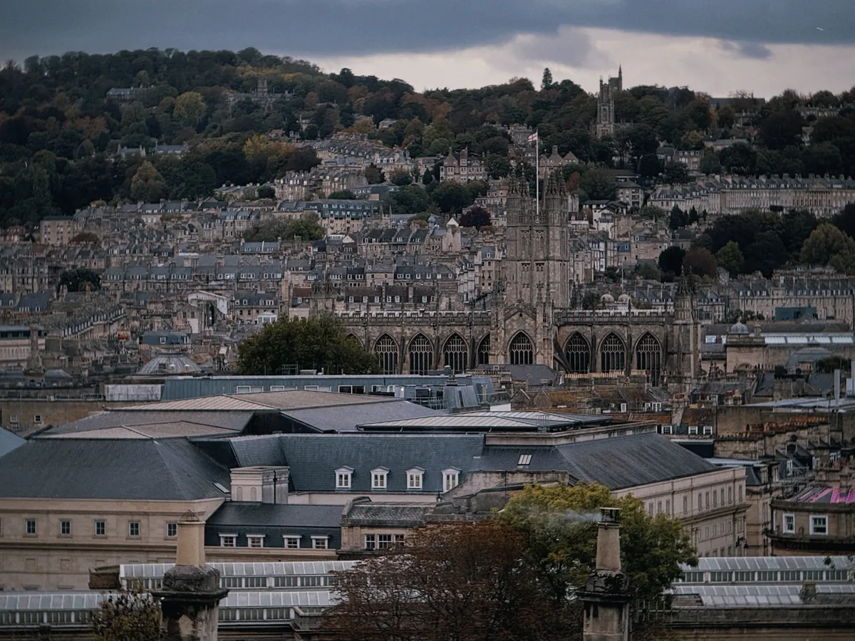 A view across the rooftops of Bath on an overcast day, with the Abbey rising among rows of honey-stone terraces and wooded hills behind