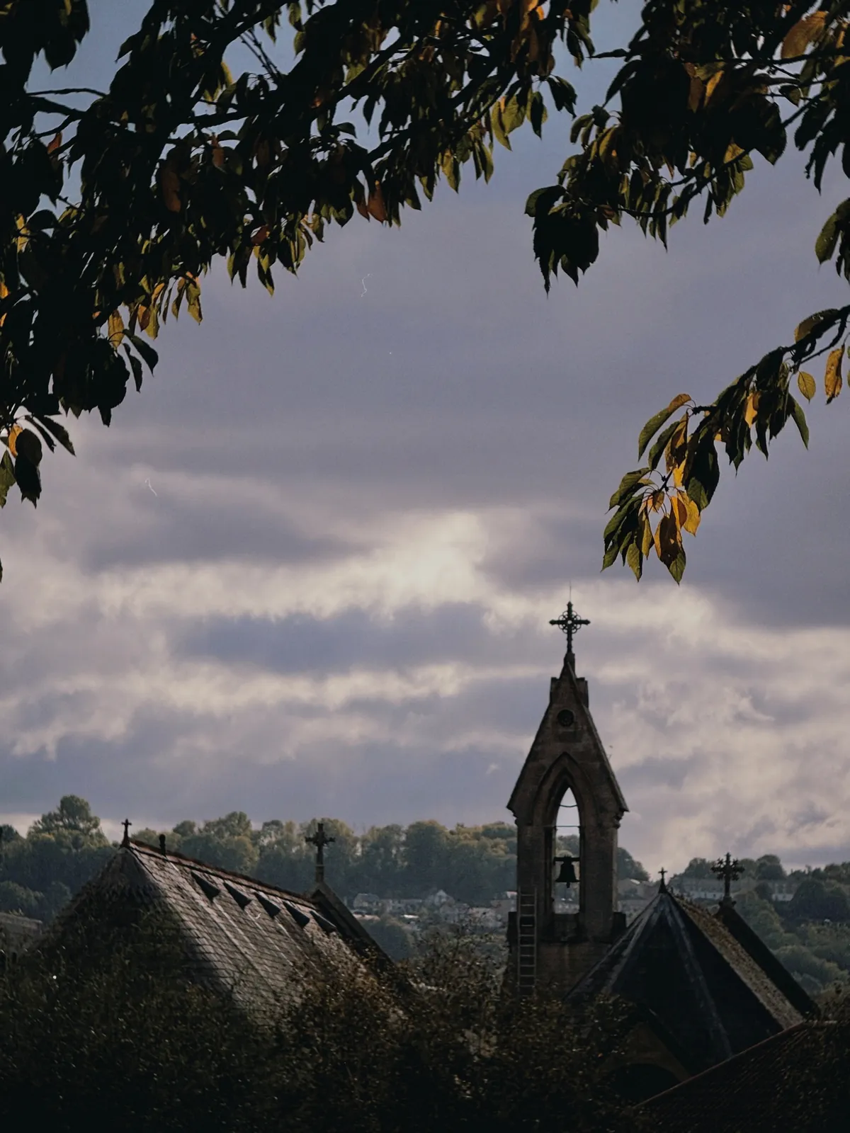The silhouetted bell tower and slate roofs of a Gothic stone church under a moody grey sky, framed by overhanging branches with yellowing leaves and wooded hills behind