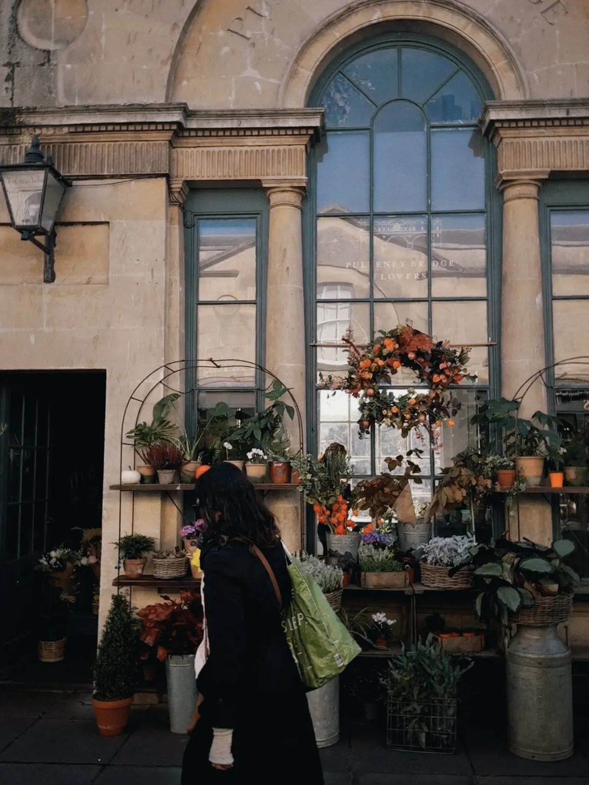 A woman with dark hair in a long black coat, carrying a green tote bag, walks past the flower shop on Pulteney Bridge