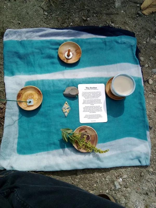 An altar cloth folded into a square on the ground. The cloth is patterned in shades of blue. At each edge sits a wooden bowl containing a symbol of the four elements: incense, candle, water, and a sprig of goldenflower. At the center of the altar are a rock, a Tessera charm depicting an anchor, and a card describing the meaning of the anchor.