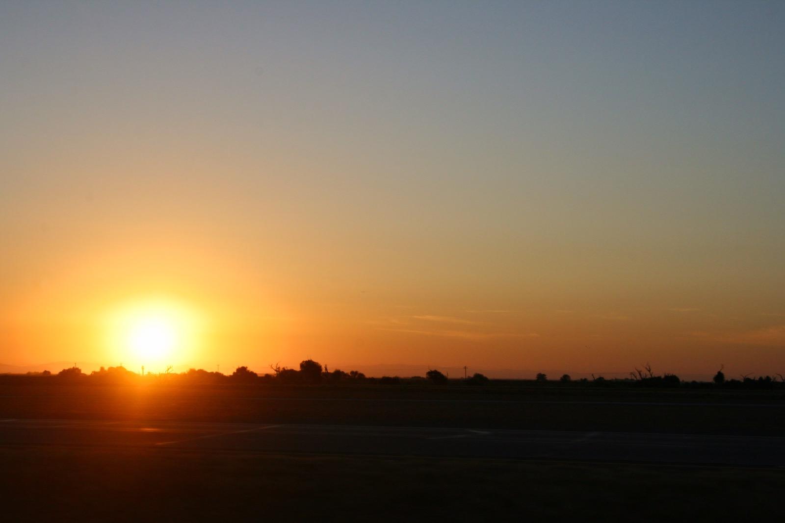 A landscape taken from a car. The sky is a rich orange gradient, and the sun hangs just over the black silhouette of the California countryside skyline.