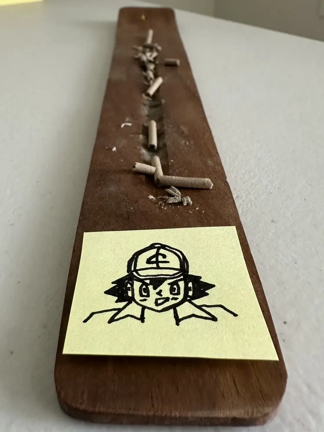 a small yellow post it note with a drawing of the lead pokemon character ash ketchum. the post it note is attached to a wooden ash catcher that people use to burn incense on. get it? rofl.