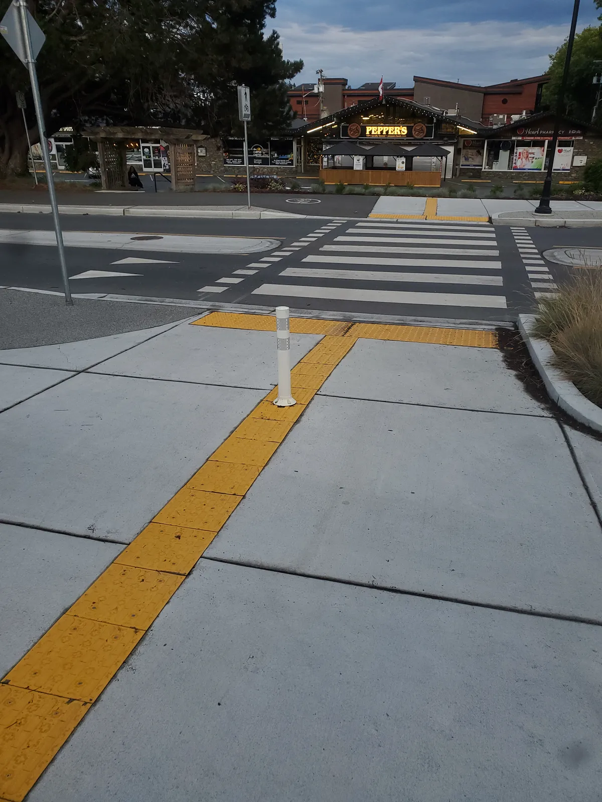 An image of the raised crosswalk + crossride over the East leg of the roundabout, with TWSIs in the foreground, a mysterious flex post in the middle of the TWSI line, and the peaked roof and lit up yellow sign for Pepper's market in the background.