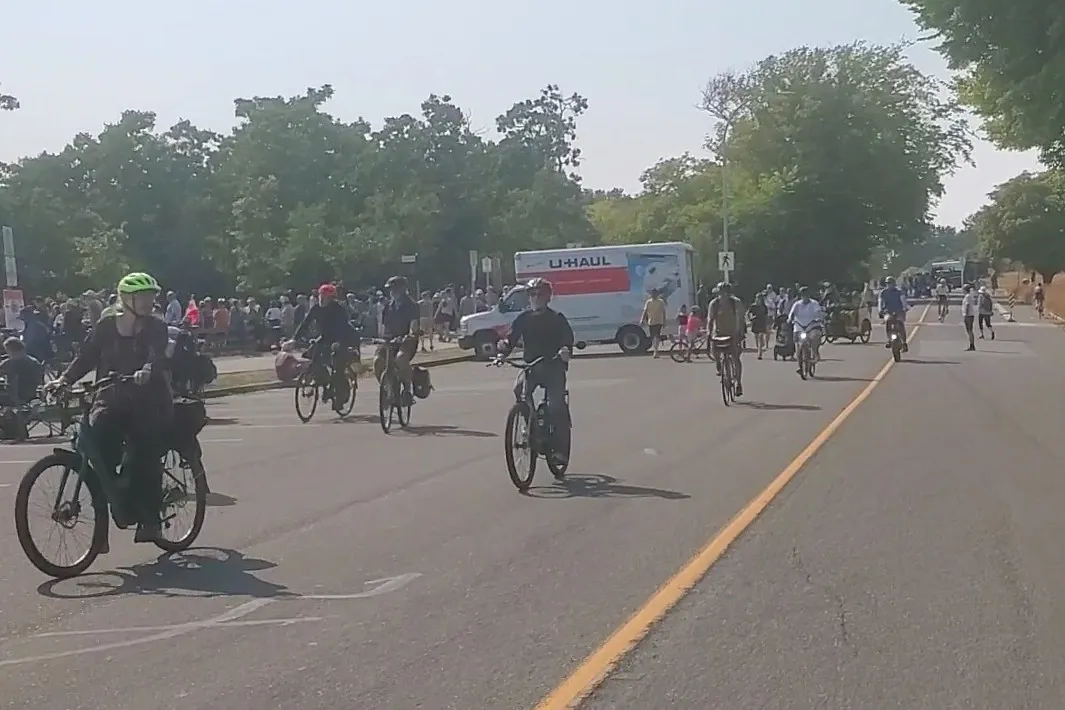 A picture taken close to the centerline on Dallas Road, looking west towards a u-haul truck in the background. In the foreground are a handful of older folks on upright ebikes, with more people cycling and walking along the road in the background.