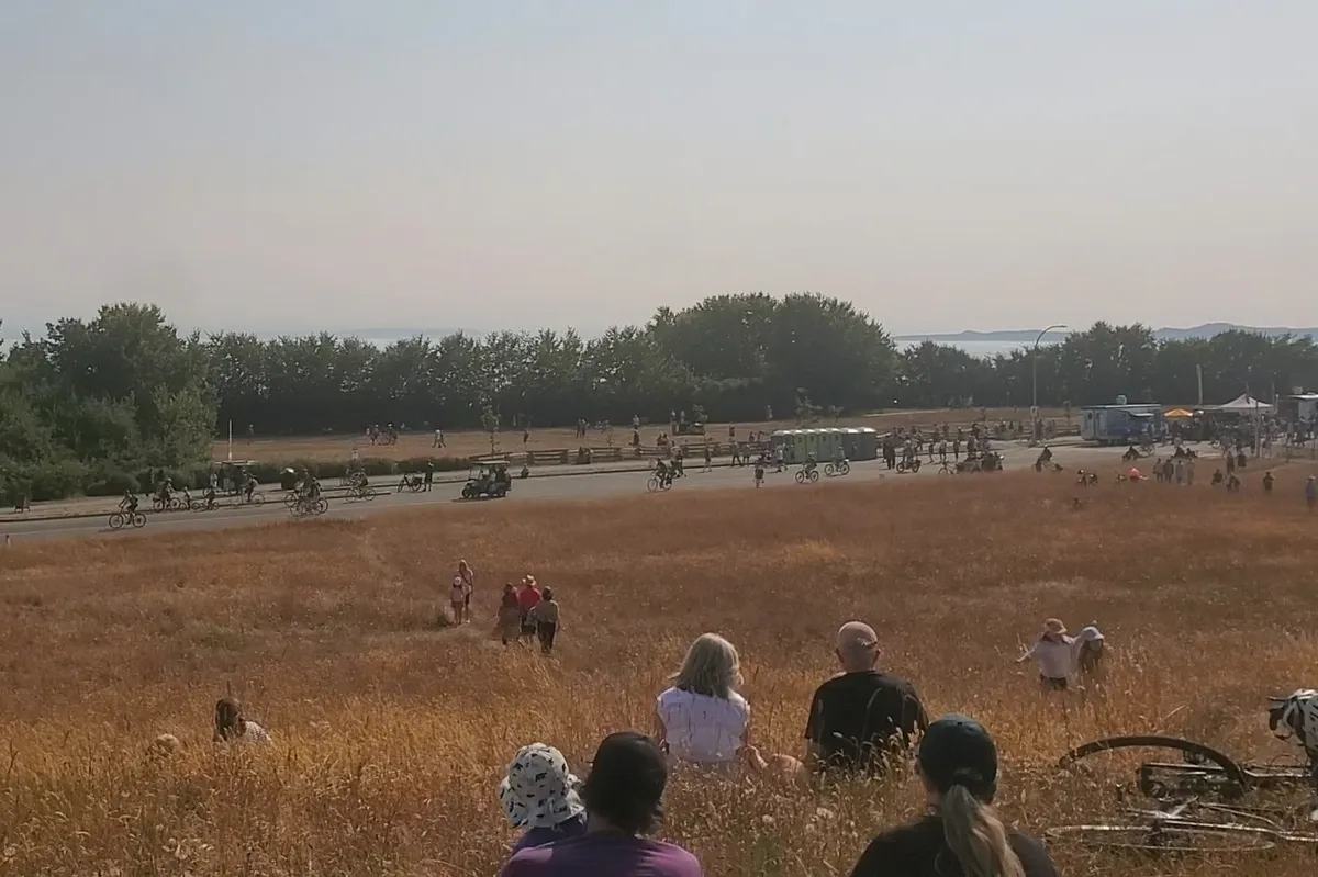 The view from up on Beacon Hill, looking down on the tall grass on the slope towards Dallas Road, with the trees, ocean and Washington's mountains visible in the distance. In the foreground are a dozen people sitting and walking on the hill, and a bike laying on its side. On Dallas Road are a few food trucks, portable toilets, and about 14 people cycling east, plus lineups near the food trucks and a golf-cart shaped 'Hey Ya' vehicle ferrying people through the event. During the entire lead-up to the air show, there were consistently people cycling along Dallas Road in both directions.
