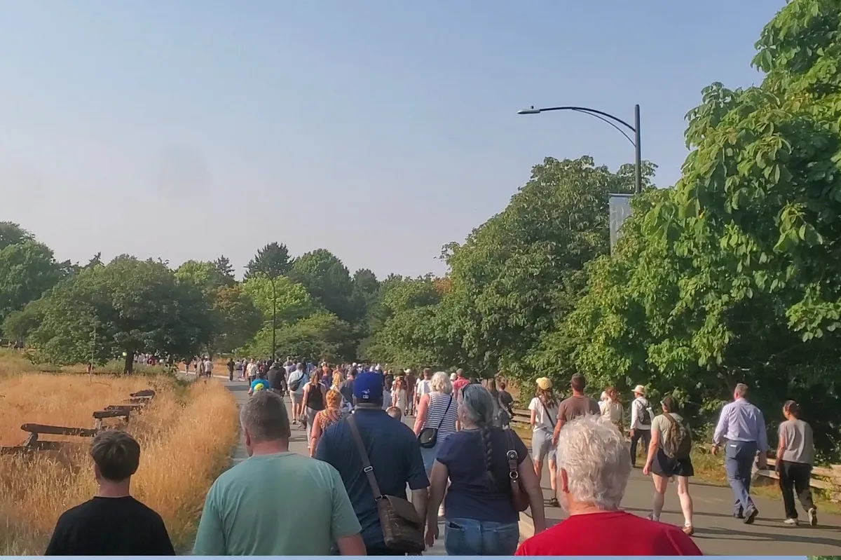 An image taken from the normally 'vehicle' side of Crescent Loop in Beacon Hill Park, a two lane wide road divided by a low concrete curb with a treeline visible on the right side and the tall grass of the Garry Oak Savannah on the left side. Along all of the road is a crowd of pedestrians heading north. Behind the camera is a clump of people waiting to bike through.