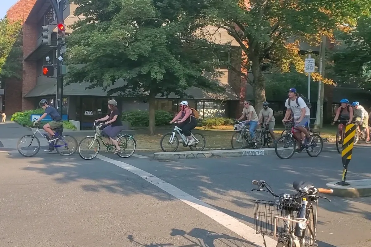 9 people on bikes riding southbound on Vancouver at Fort street. Most of them are in a line in the southbound protected bike lane, a few are using the street. This is a still from a video where a few dozen people cycle through this traffic signal, with a large number of people coming from further down Vancouver to repeat the same events during the next light cycle.