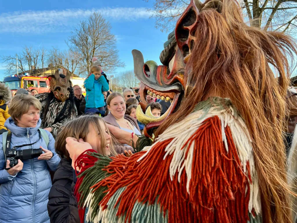 Eine große, zottelige Teufelsmaske mit Zähnen und Hörnern interagiert mit lachenden Zuschauern am Straßenrand.