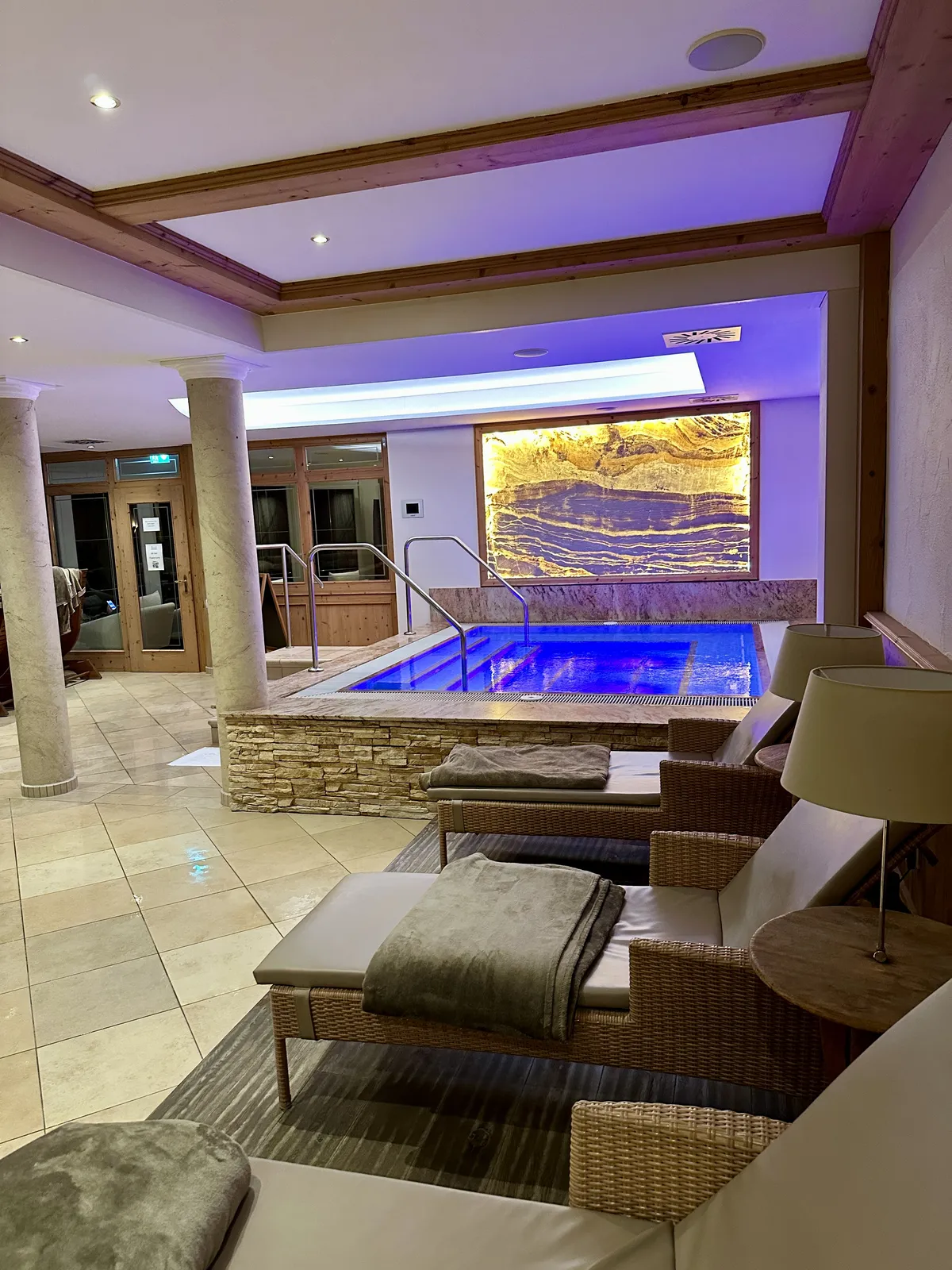 Ein moderner Wellnessbereich mit einem kleinen Pool, umgeben von Steinwänden und beleuchtet mit sanftem, violettem Licht. Im Vordergrund sind Liegen mit grauen Decken und ein Tisch mit einer Lampe zu sehen.