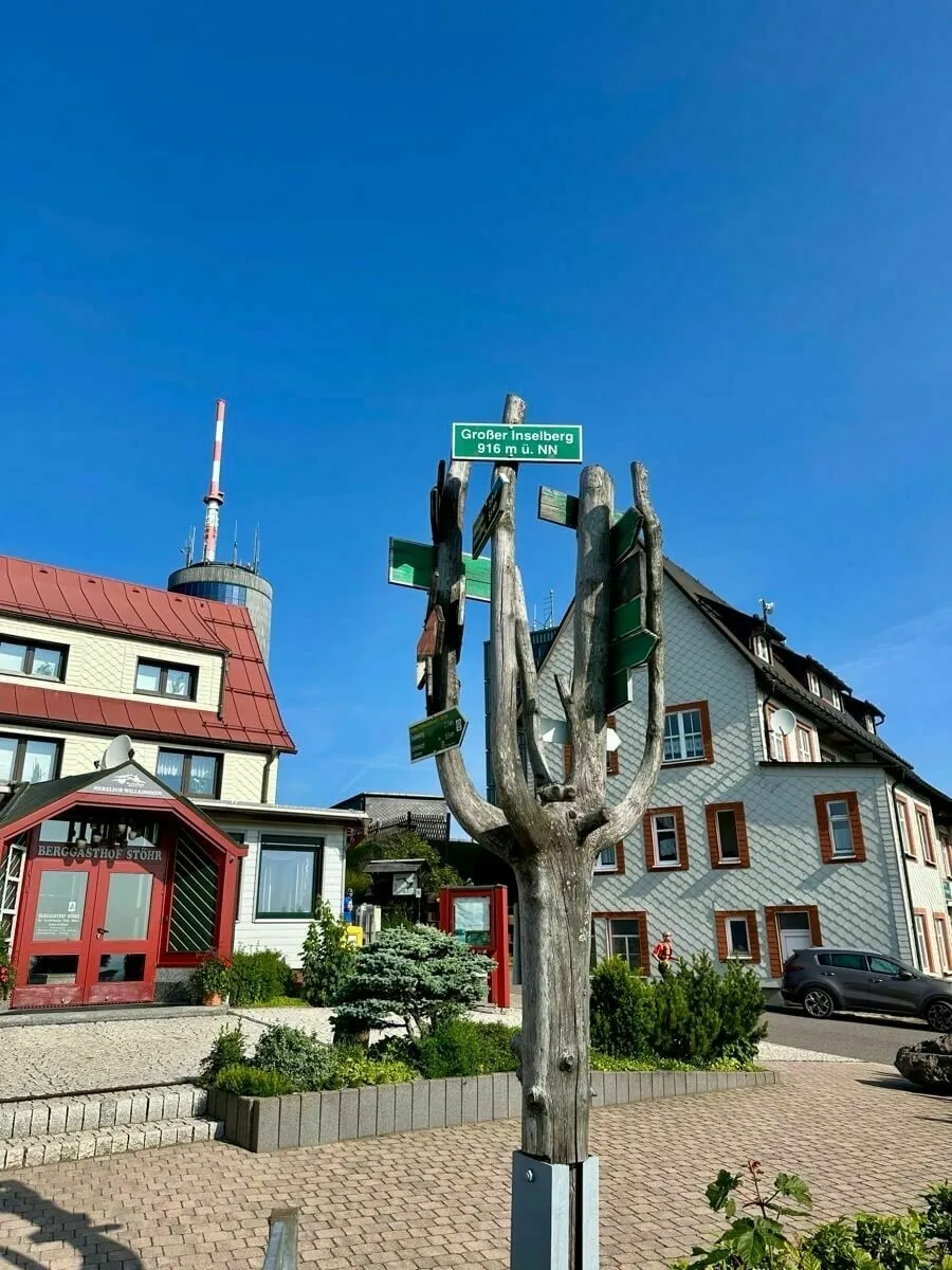 Ein kunstvoller Wegweiser in Form eines kleinen Baumes mit mehreren Schildern steht vor traditionellen Gebäuden unter einem klaren blauen Himmel.