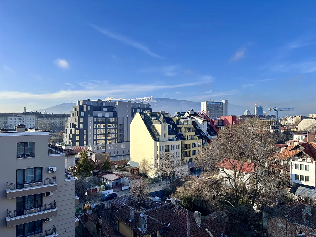 Eine Ansicht von Sofia, zeigt eine Vielzahl von Gebäuden mit unterschiedlichen Architekturstilen, darunter moderne und traditionelle Häuser. Im Hintergrund sind schneebedeckte Berge unter einem klaren blauen Himmel sichtbar.