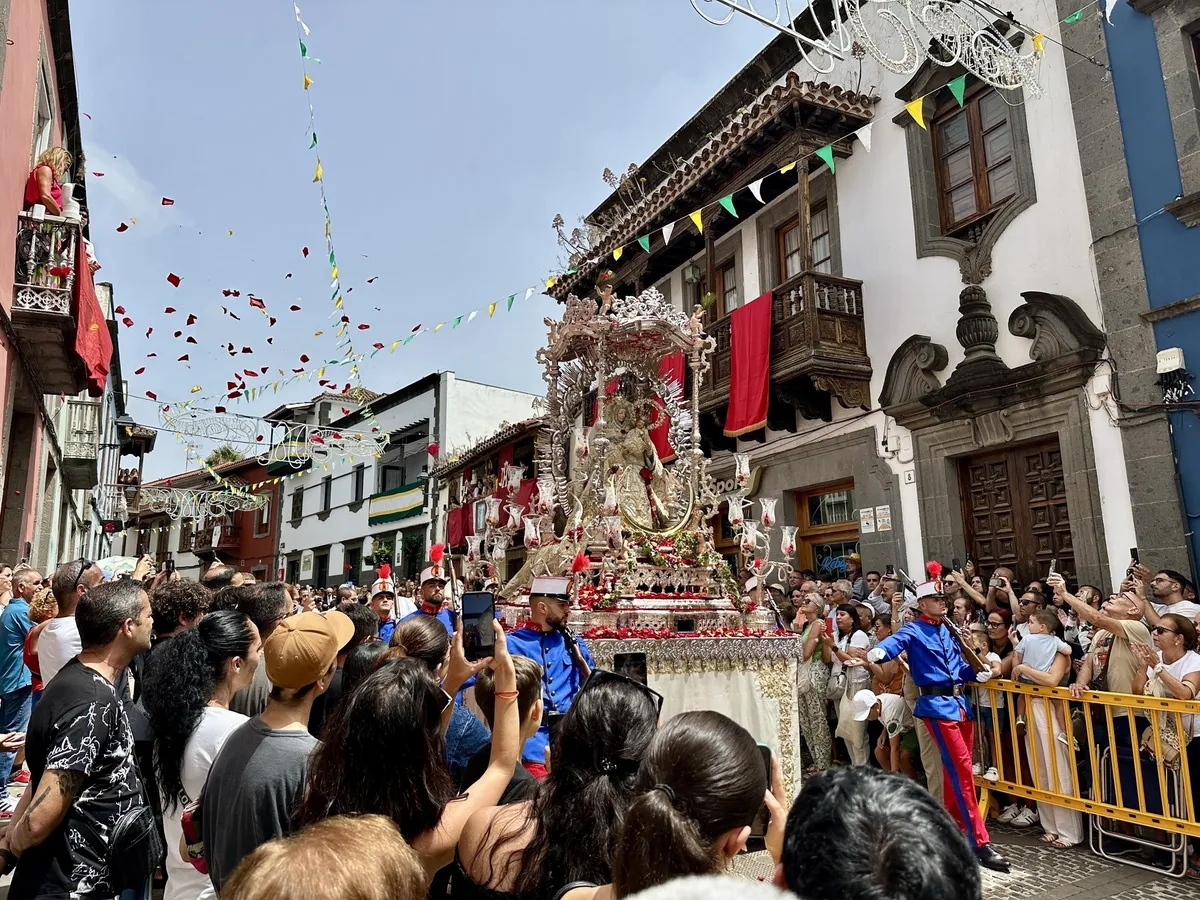 Menschenmengen versammeln sich um einen festlichen Umzug mit einem aufwendig geschmückten Wagen auf einer belebten Straße, während Blumenblätter von oben fallen.