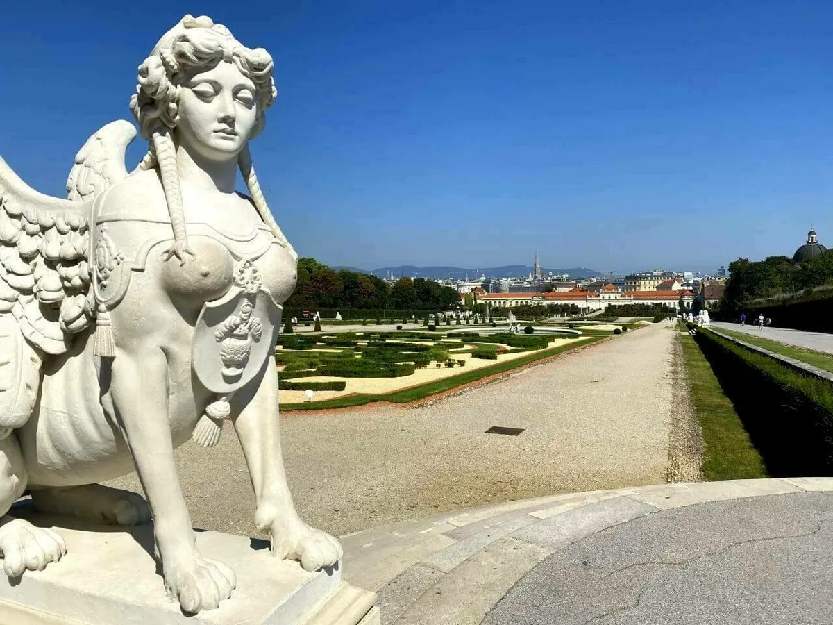 Eine Sphinx-Statue im Garten des Belvedere im Vordergrund, mit einem weiten Blick über die geometrisch angelegten Barockgärten und die Skyline von Wien im Hintergrund.