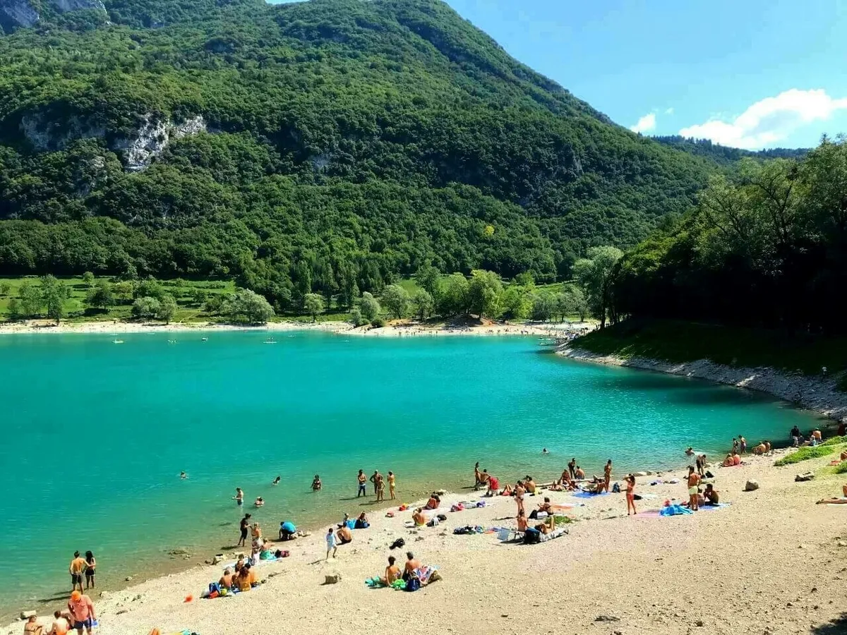 Der Lago di Tenno, ein wunderschöner Bergsee mit hellem, türkisfarbenem Wasser und einem Sandstrand. Im Hintergrund erheben sich grüne, bewaldete Berge. Viele Menschen genießen das sonnige Wetter am Strand und im Wasser.