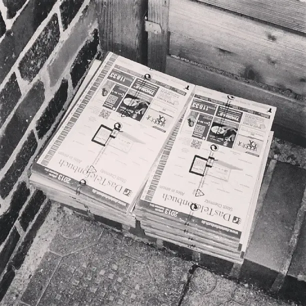 Stapel von Zeitungen oder Prospekten, die ordentlich nebeneinander auf einem Pflastersteinboden liegen, umgeben von einer Mauer und einem Holzzaun. Die Szene ist in Schwarz-Weiß gehalten.