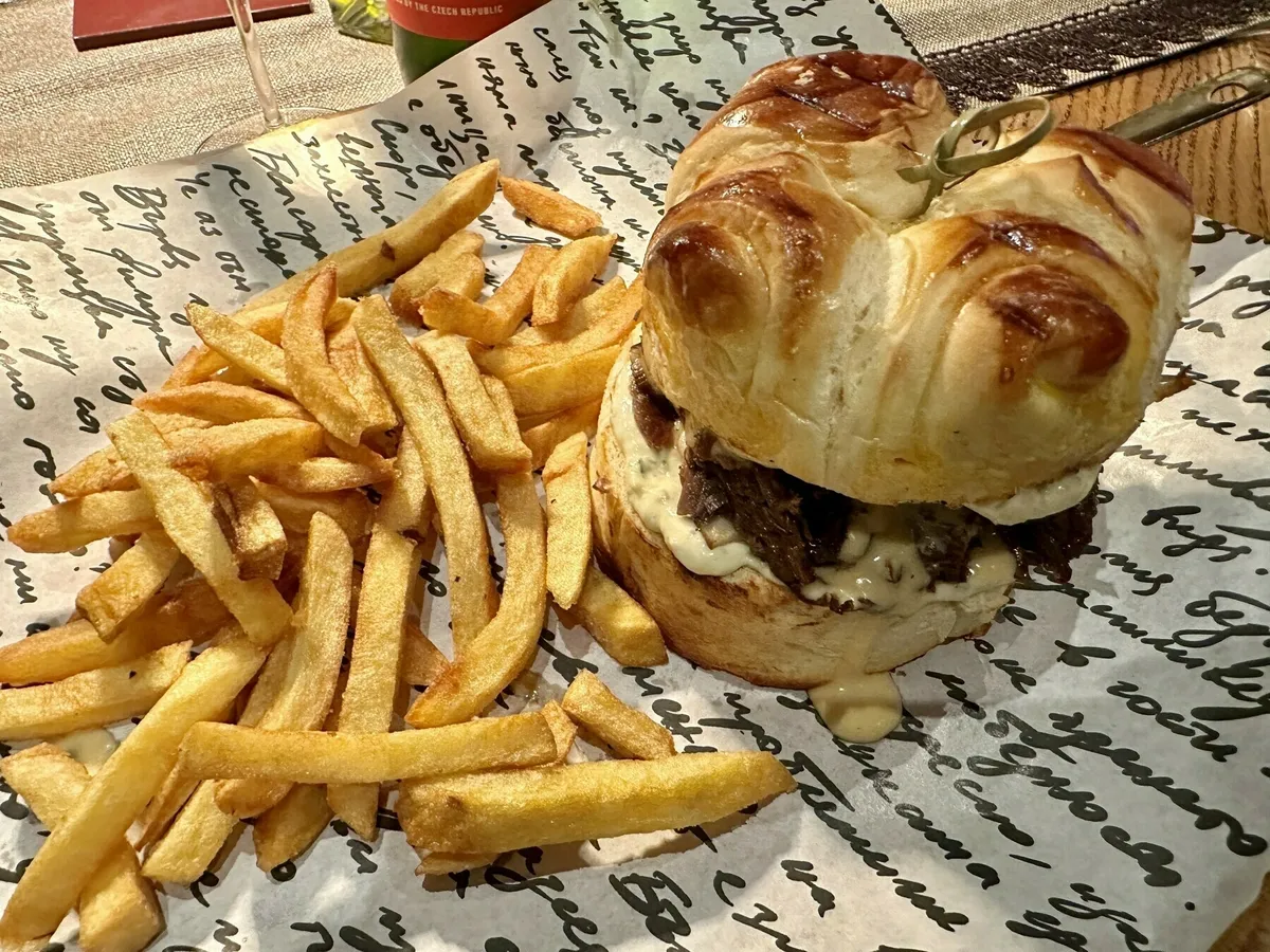 Ein großer Burger, serviert auf einem Brioche-Bun mit geschmortem Fleisch und Käsesauce, daneben ein Berg goldener Pommes Frites, alles auf bedrucktem Papier.