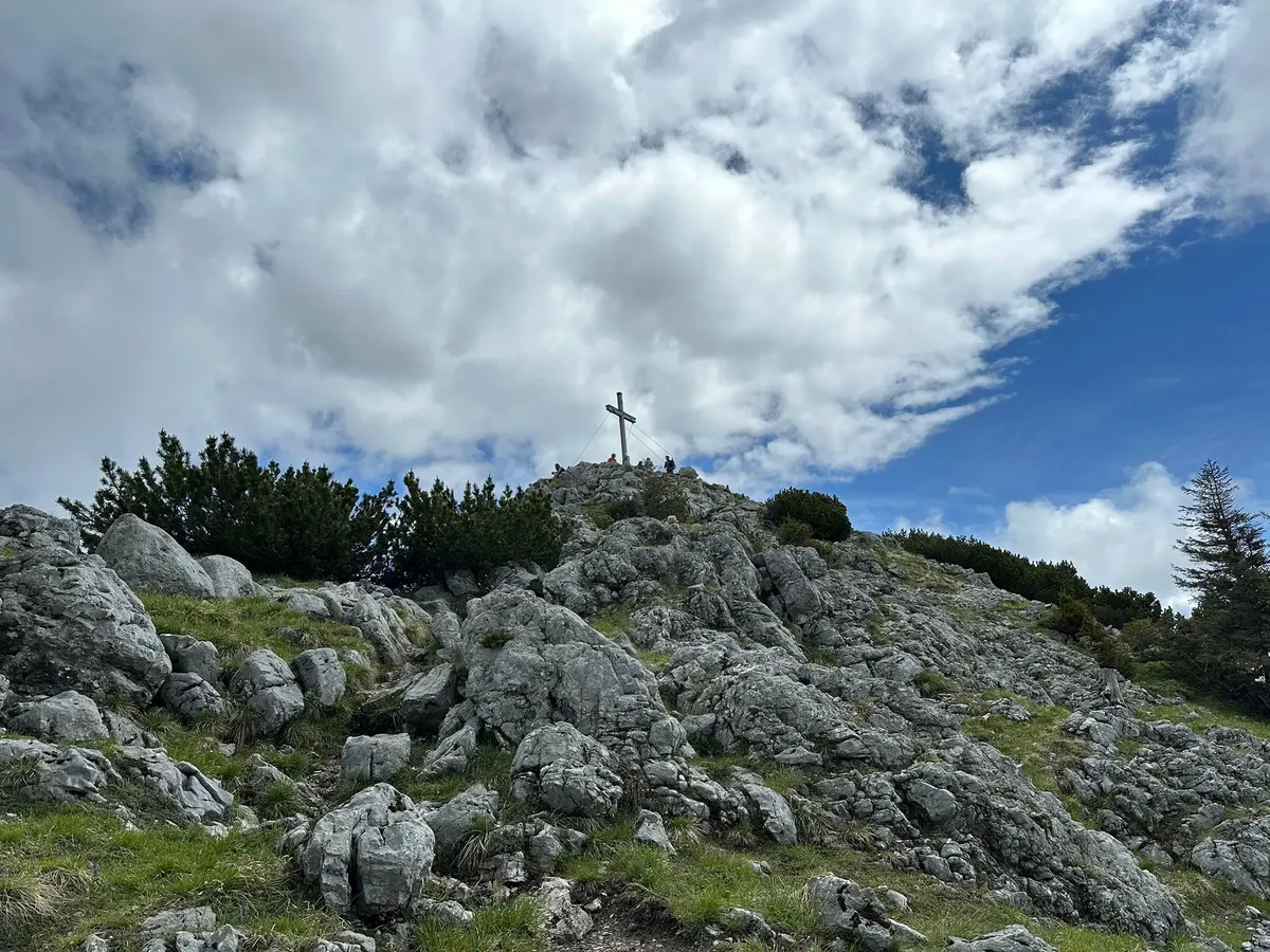 Ein Gipfelkreuz steht auf einem felsigen Hügel unter einem teilweise bewölkten Himmel.