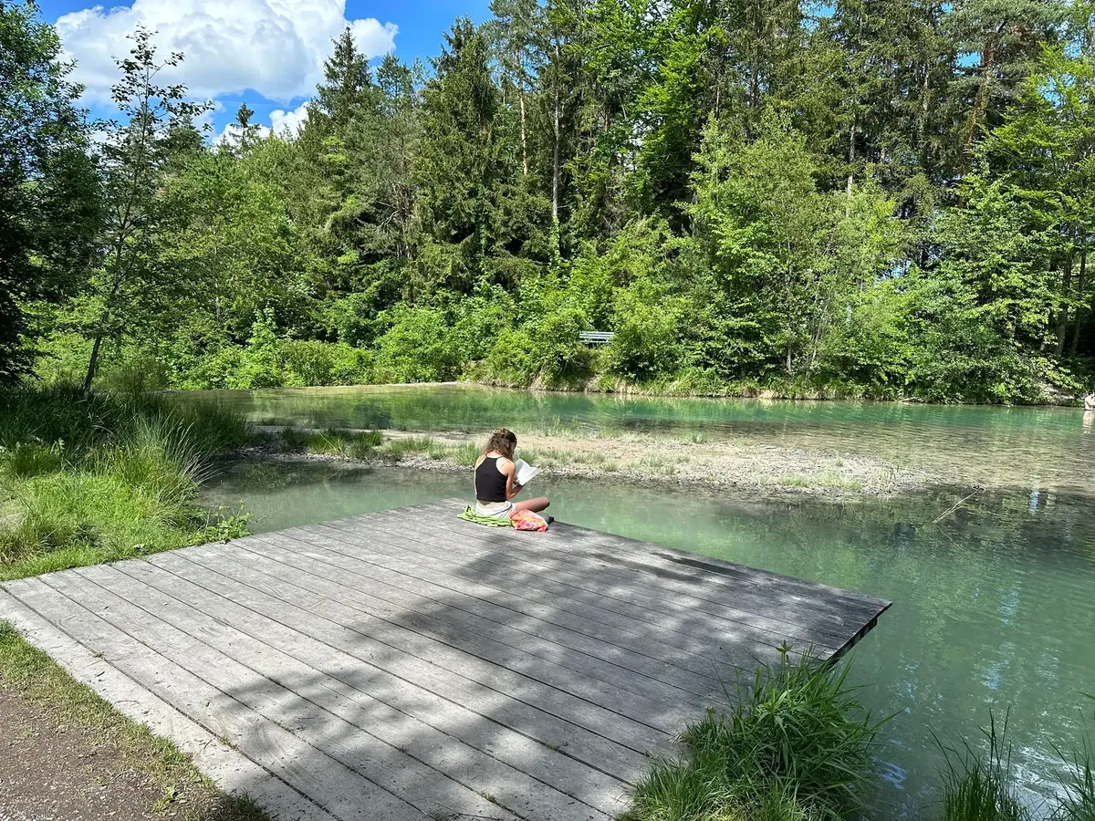 Eine Person sitzt auf einem Holzsteg an einem ruhigen, von Bäumen umgebenen See.