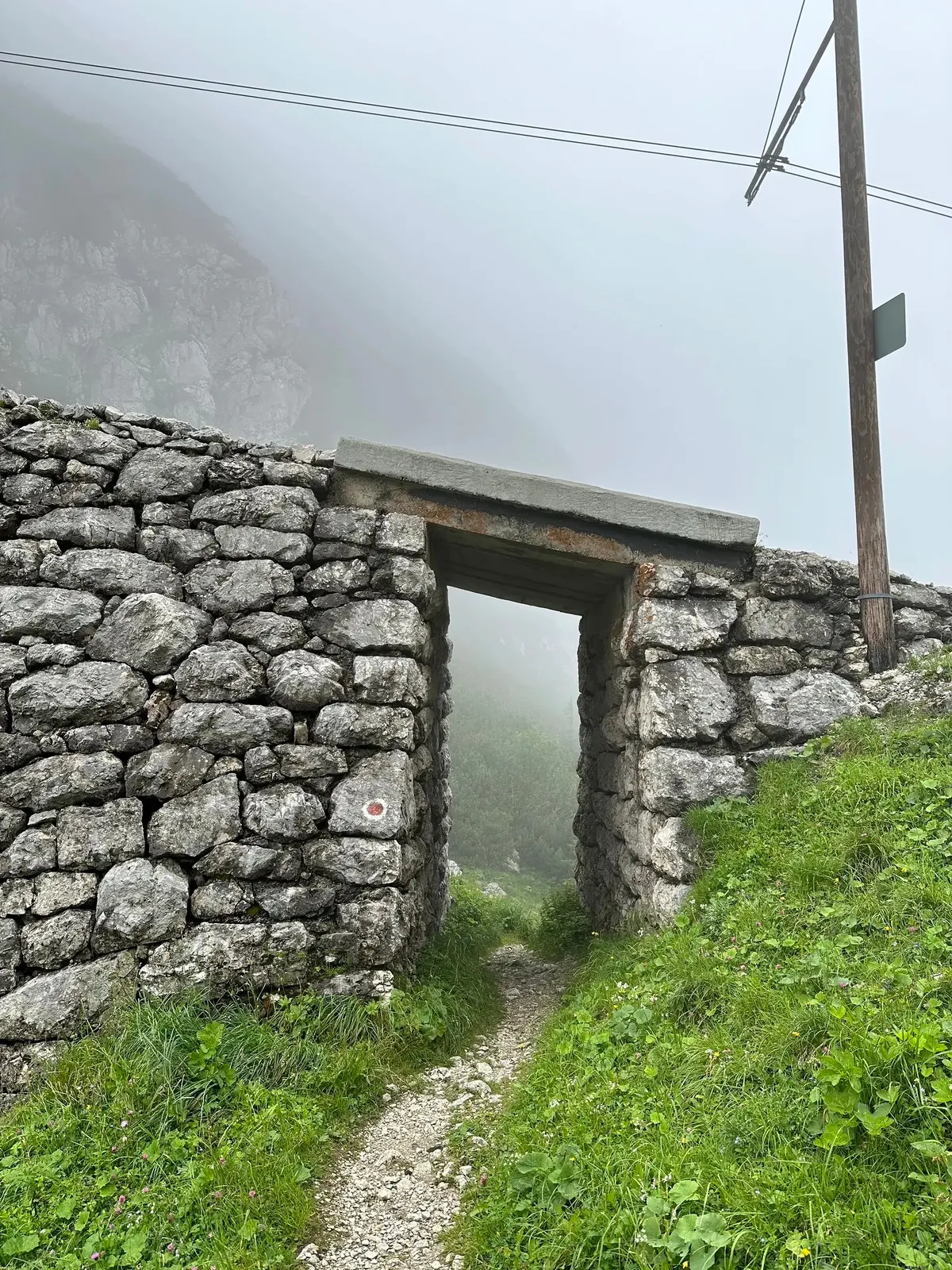 Ein schmaler Pfad führt durch eine Steinmauer in einer nebligen, bergigen Landschaft.