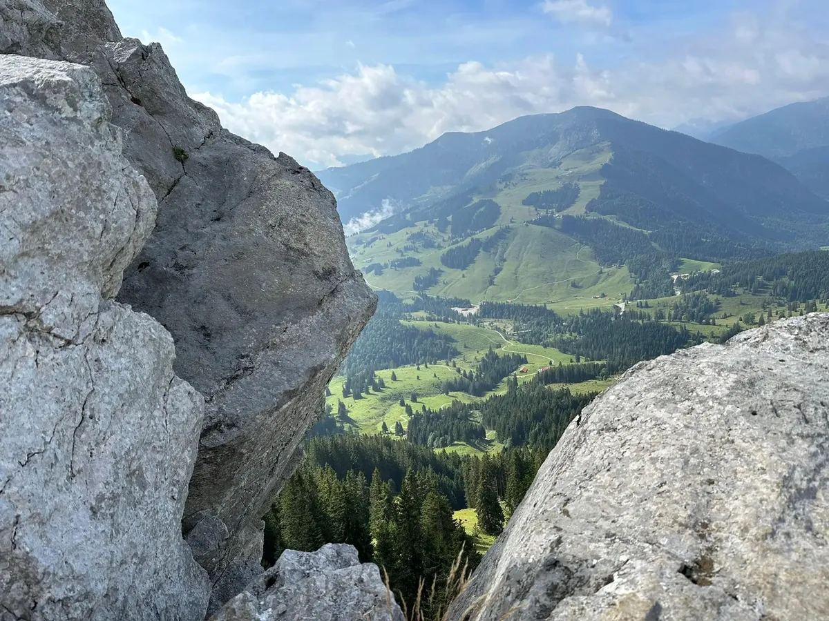 Ein Ausblick durch zwei große, graue Felsbrocken hindurch auf ein tiefes, grünes Alpental mit Wiesen, Wäldern und Wanderwegen. Die umliegenden Berge sind sanft und mit viel Grün bedeckt.