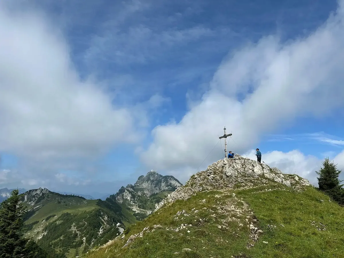 Eine weite Aufnahme eines felsigen, grasbewachsenen Bergrückens mit einem Gipfelkreuz in der Mitte. Zwei Personen stehen neben dem Kreuz und blicken in die Ferne auf die umliegenden, zerklüfteten Berge. Der Himmel ist blau mit großen, weißen Wolken.