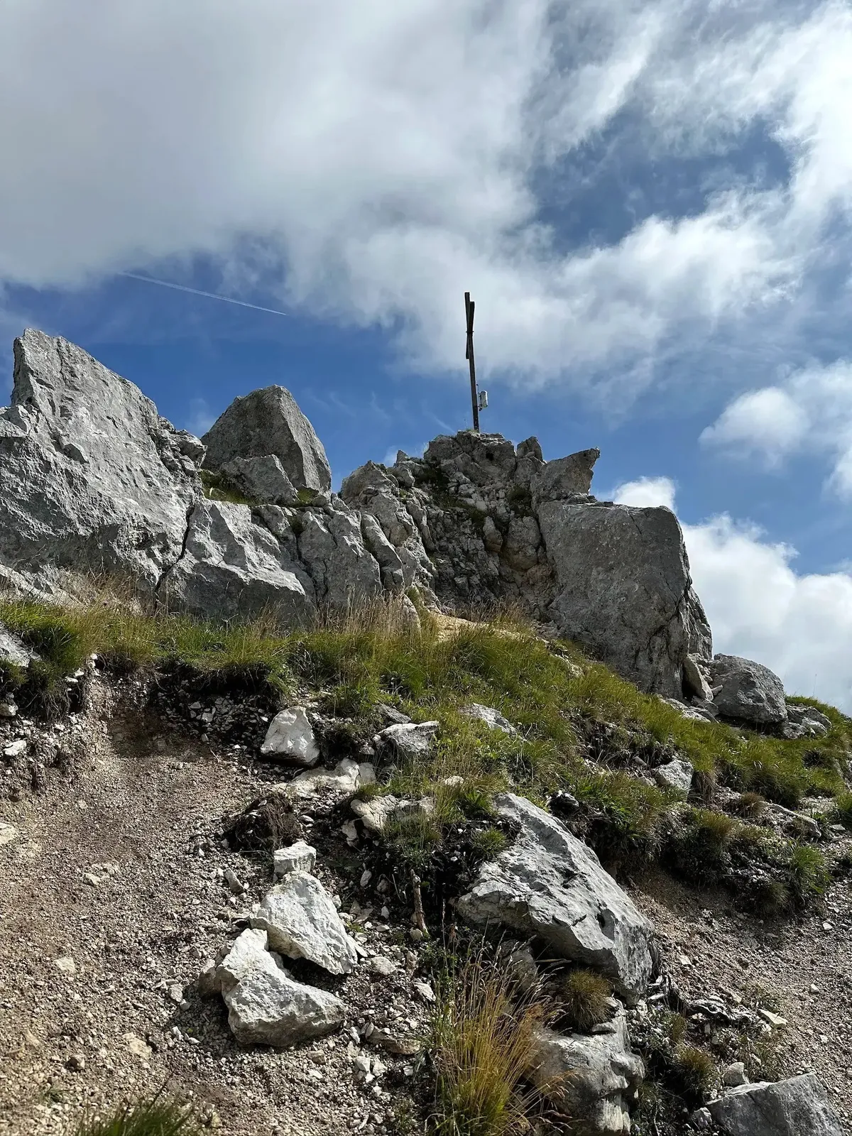 Ein Gipfelkreuz steht auf einem Haufen großer, grauer Felsbrocken und Schutt. Im Vordergrund wachsen trockenes Gras und kleinere Pflanzen. Der Himmel ist blau mit weißen Wolken.