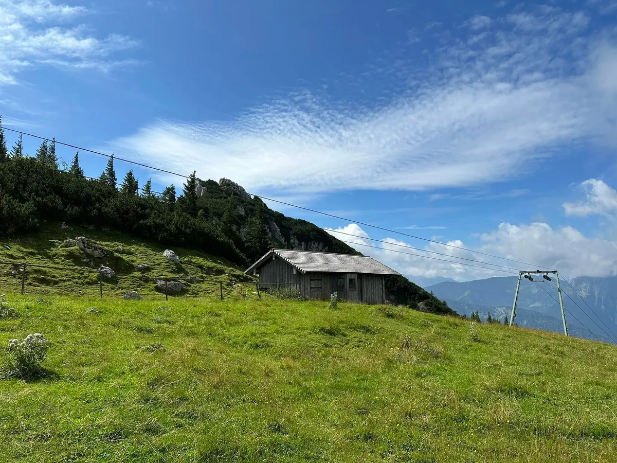 Eine kleine, rustikale Holzhütte mit einem Blechdach steht auf einer grünen Bergwiese. Rechts führt eine Seilbahnkonstruktion den Berg hinauf. Im Hintergrund sind weitere Berge und der blaue Himmel zu sehen.