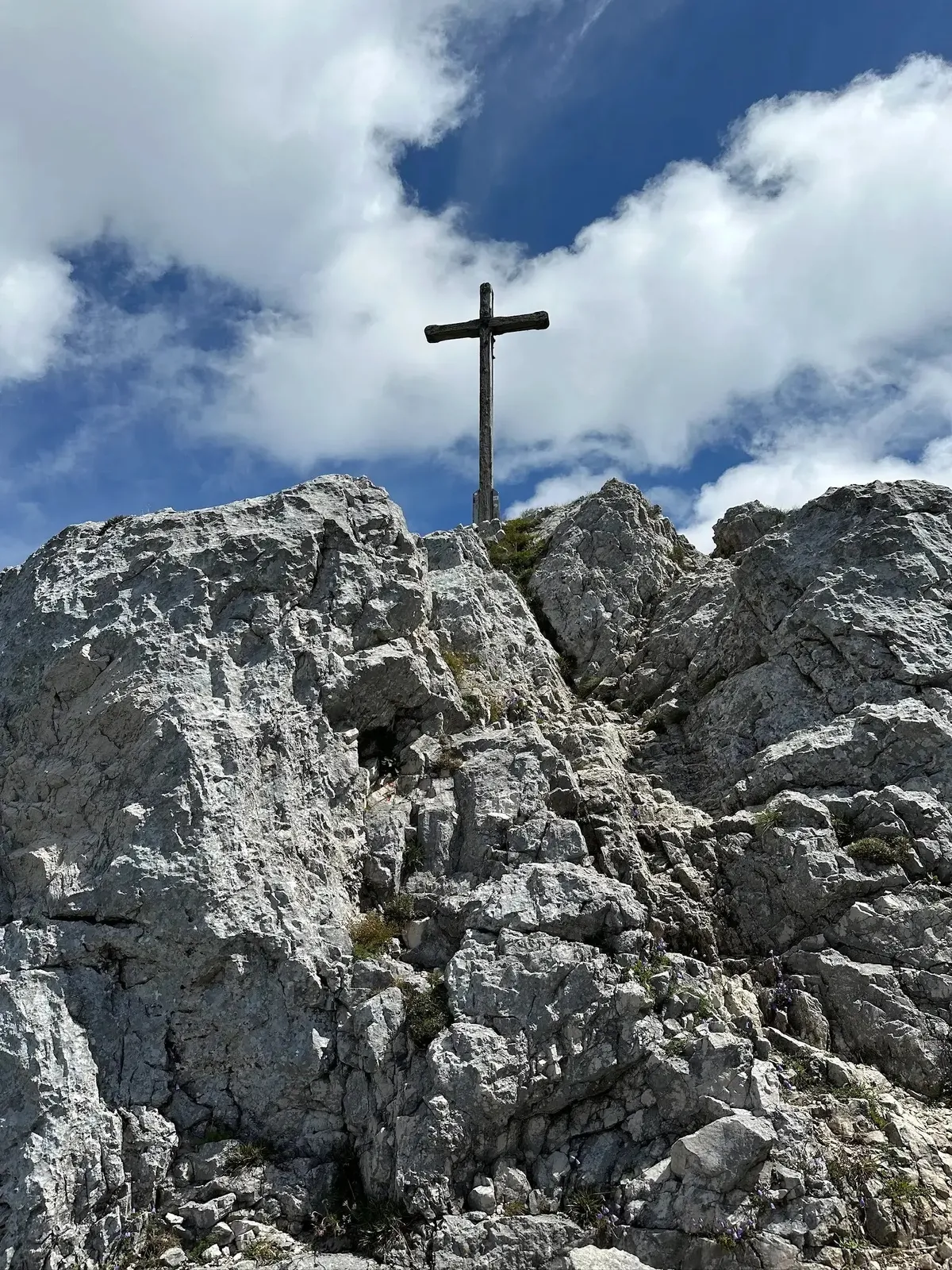 Ein robustes, dunkles Gipfelkreuz auf einem zerklüfteten, felsigen Berggipfel, aufgenommen von unten. Der Himmel ist hellblau mit dramatischen weißen Cumuluswolken.