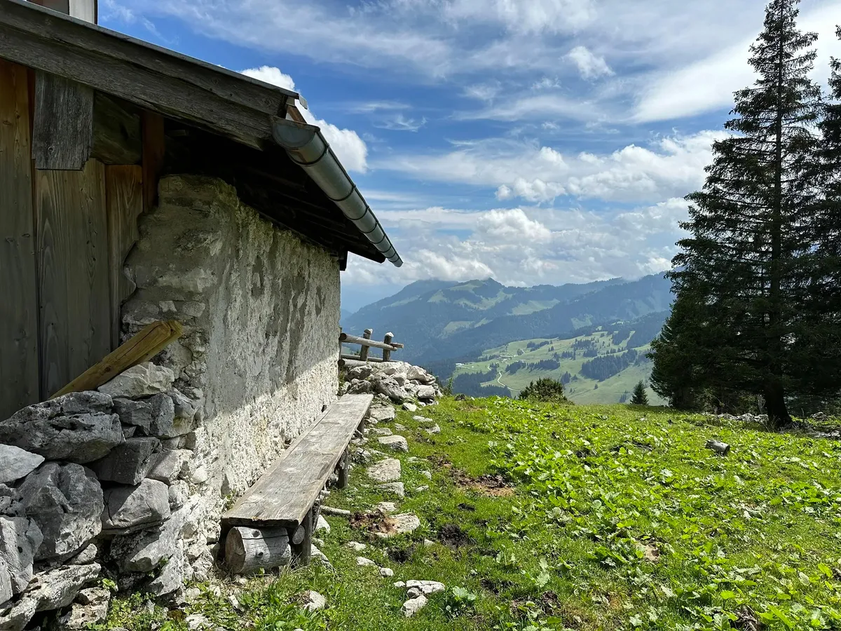Eine Nahaufnahme der steinernen Außenwand einer Almhütte mit einem hölzernen Vordach und einer einfachen Holzbank. Von der erhöhten Position bietet sich ein weiter Blick über grüne, bewaldete Berghänge.