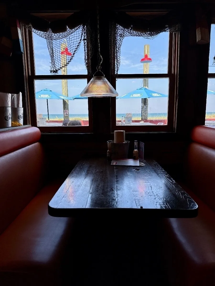 Innenansicht einer holzgetäfelten Restaurant-Nische mit gepolsterten orangefarbenen Sitzbänken und einem dunklen Holztisch. Durch die Fenster sieht man Sonnenschirme, den Strand und das Meer. Ein aufgehängtes Netz und eine Lampe zieren den oberen Bereich der Fenster.